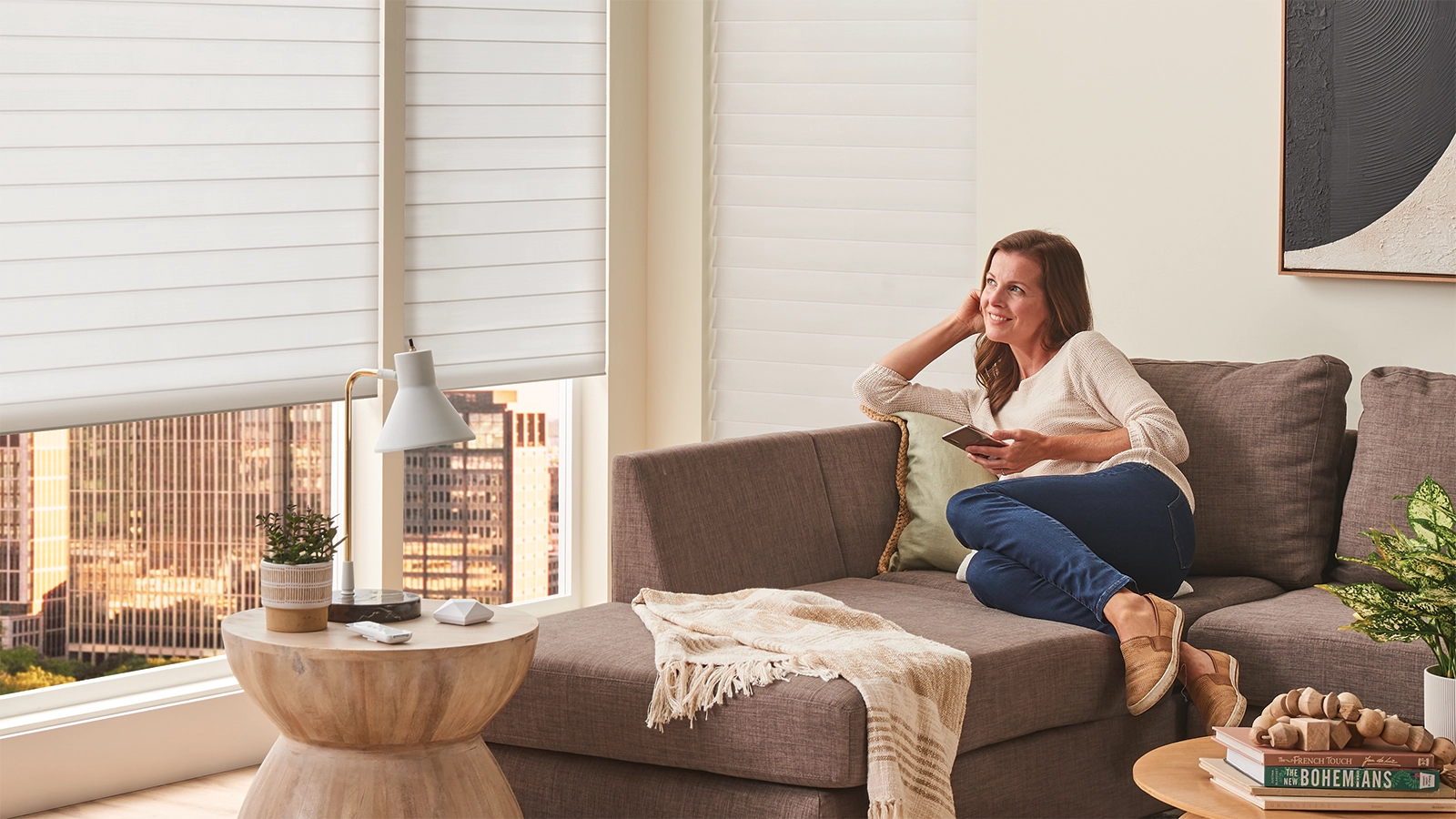 Woman sitting on her sofa using the Alta BLISS Automation app on her phone to open the shades in her living room.